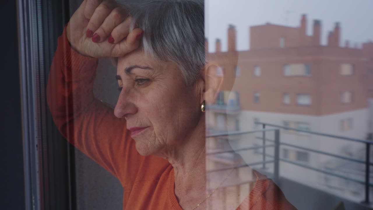 Mujer mayor pensativa mirando por la ventana con reflejo urbano.