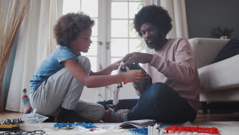 father sitting on the floor at home helping his pre teen son construct a kit toy, close up, low angle