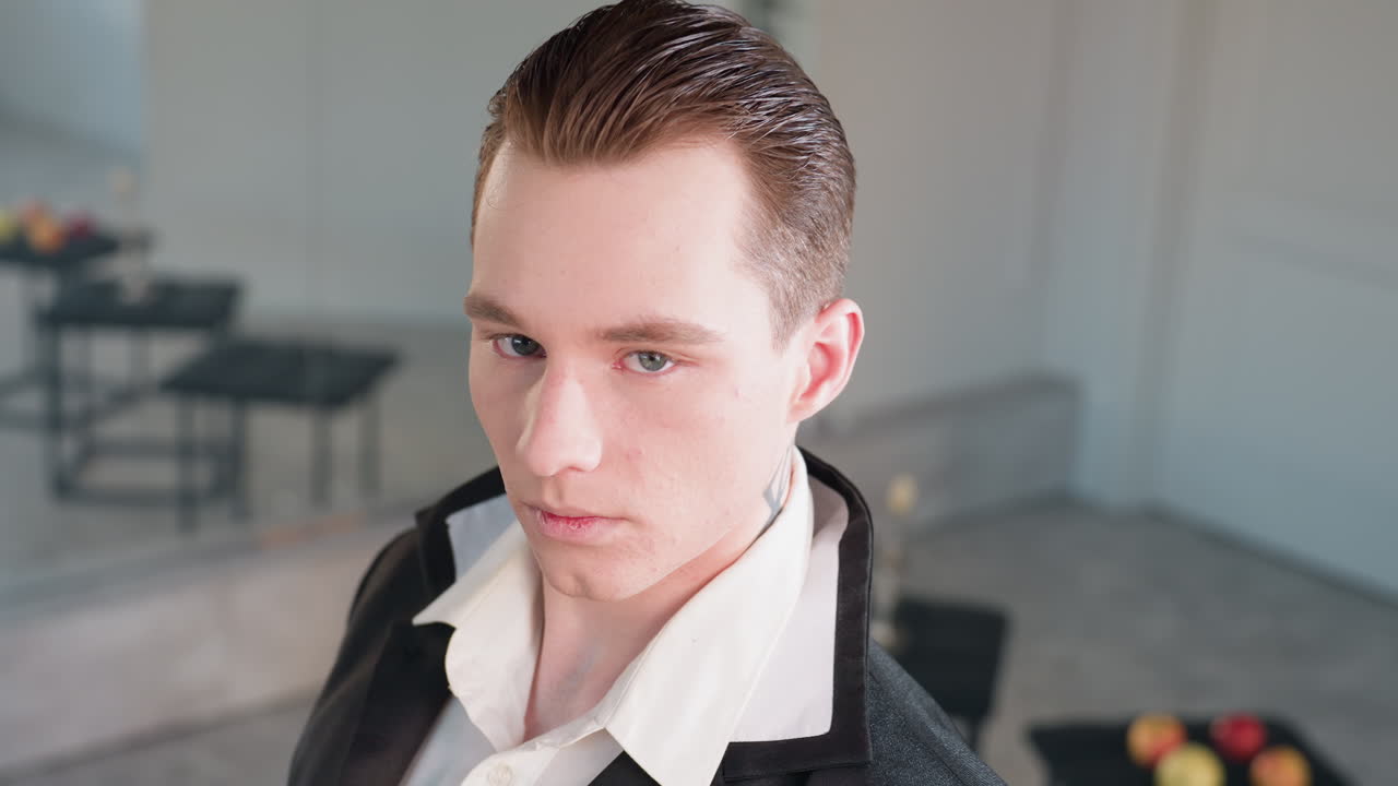 stunning young man with slicked hair and neck tattoo gazes elegantly across modern studio interior, three stools behind with blurred apples and candle stand, large wall mirror