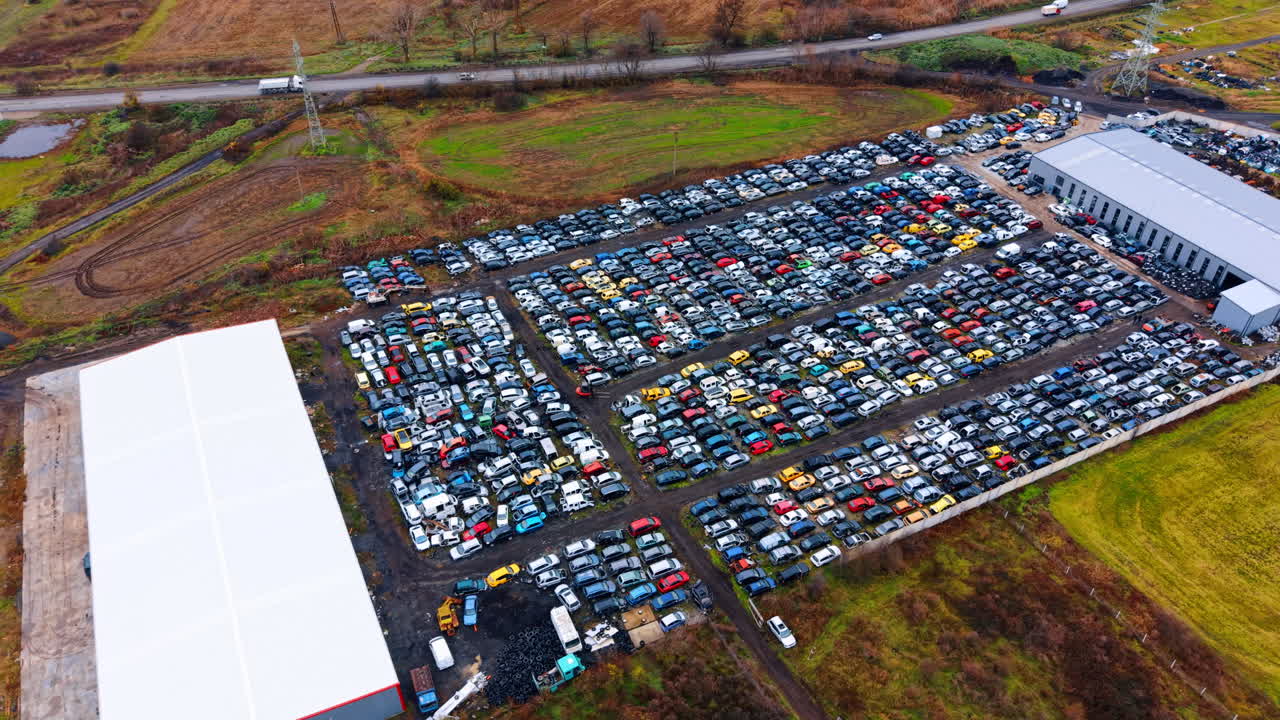 Vista aérea panorámica de un desguace automotriz y campos circundantes. Toma de ángulo alto que muestra una vasta colección de coches chatarra, una nave industrial y caminos rurales.