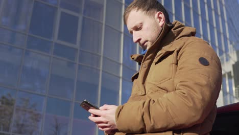 young man sitting by the car, using his smart phone, modern building with big windows at the background