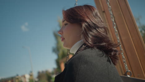 close-up of young lady swinging outdoors, hair swaying gently with breeze, with vibrant greenery, blurred urban buildings, and bright blue sky