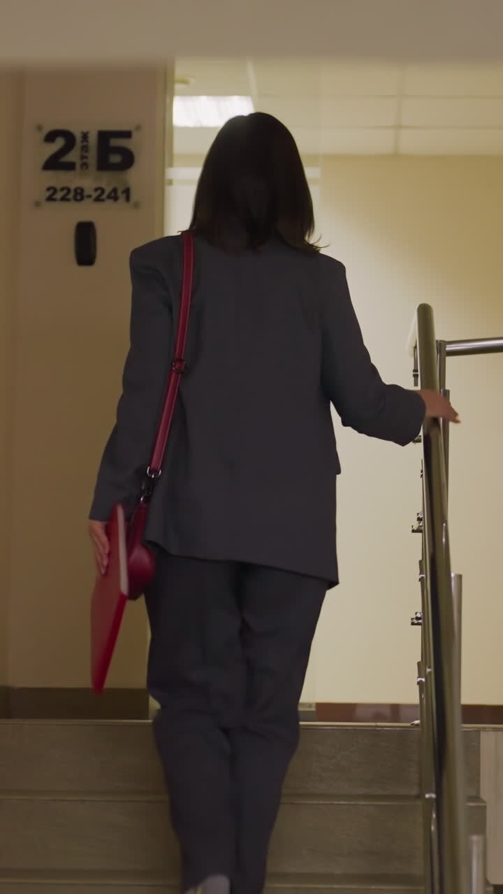 Forretningskvinde går op ad trappen med rød læderhåndtaske og notesbog på vej til arbejde. Selvsikker og professionel i et kontormiljø, der symboliserer motivation og fokus i karrieren.