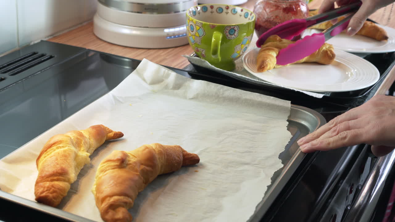 French croissants being put on white breakfast plates with jam and coffee-tea on tray by female mothers hand using pink kitchen tongs