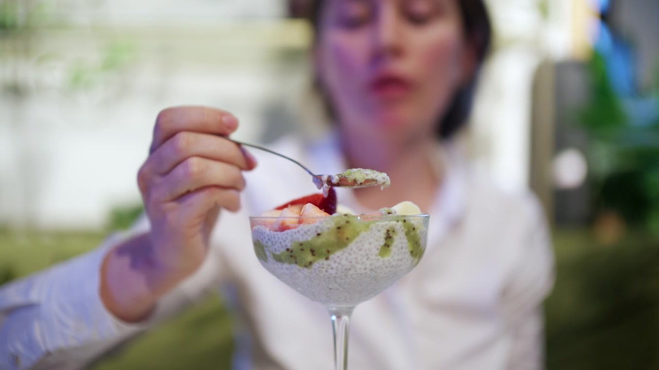 Una donna tiene un cucchiaio di budino di chia davanti a un bicchiere da dessert riempito con budino di semi di chia bianchi, purea di kiwi verde, fragole a fette e banana.