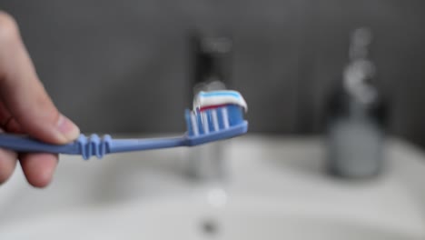 primer plano de manos caucásicas apretando pasta de dientes en un cepillo azul frente al lavabo blanco