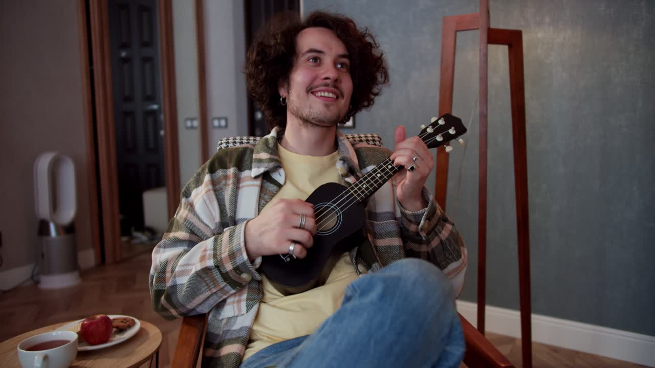 feliz chico moreno en una camisa a cuadros con cabello rizado toca el ukulele y se sienta en una silla en un acogedor apartamento moderno