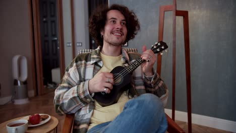 Happy-brunette-guy-in-a-checkered-shirt-with-curly-hair-plays-the-ukulele-and-sits-on-a-chair-in-a-cozy-modern-apartment