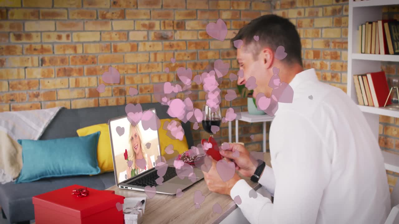iconos de corazón rosado sobre hombre caucásico sosteniendo un cartel en forma de corazón mientras hace una videollamada en una computadora portátil