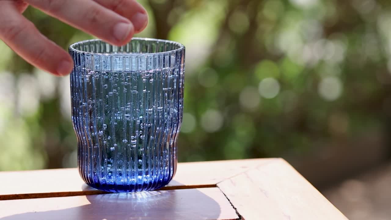 A hand approaches a textured blue glass on a sunlit wooden table, surrounded by a blurred green background.