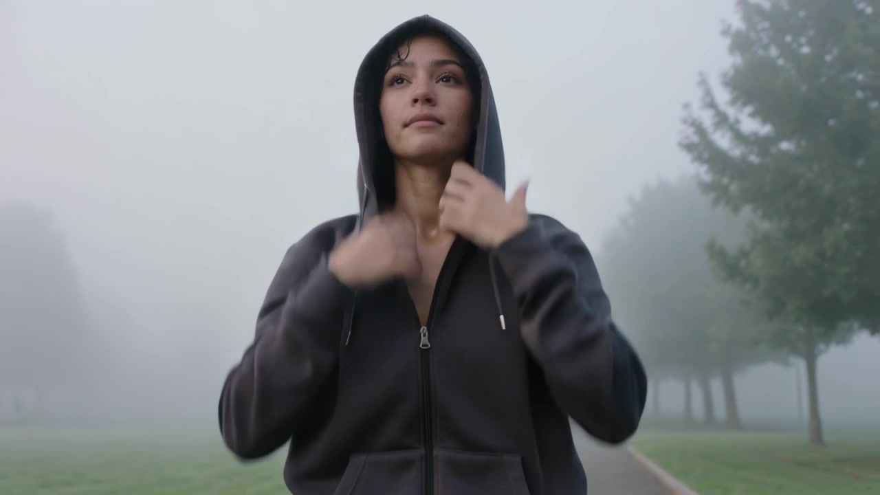 Donna che si allunga, tirando su e chiudendo la cerniera di una felpa scura con cappuccio su un sentiero nebbioso del parco, preparandosi per una corsa.