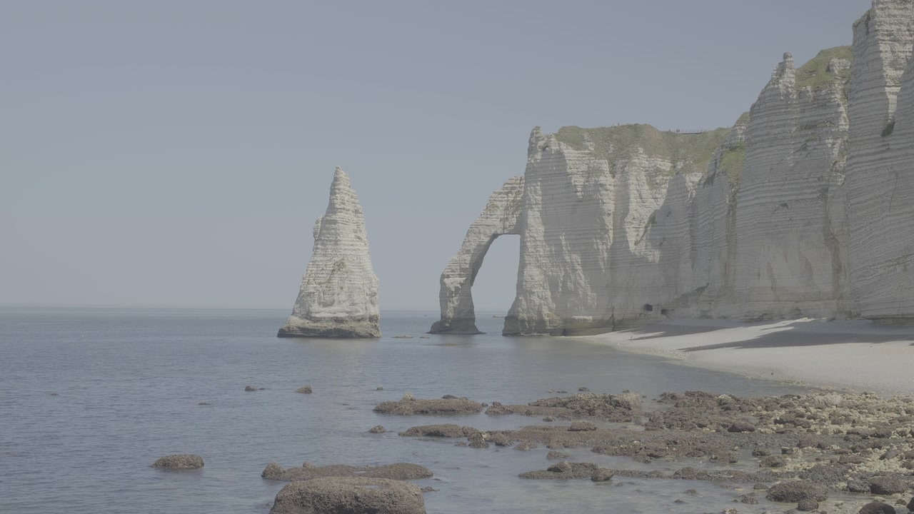 Etretat tebeşir kayalıklarının sakin mavi bir denize karşı, açık gökyüzü altında doğal güzelliği sergileyen, nefes kesen geniş çekimi; sinematik kalite, dinamik hareket, atmosferik aydınlatma LOG