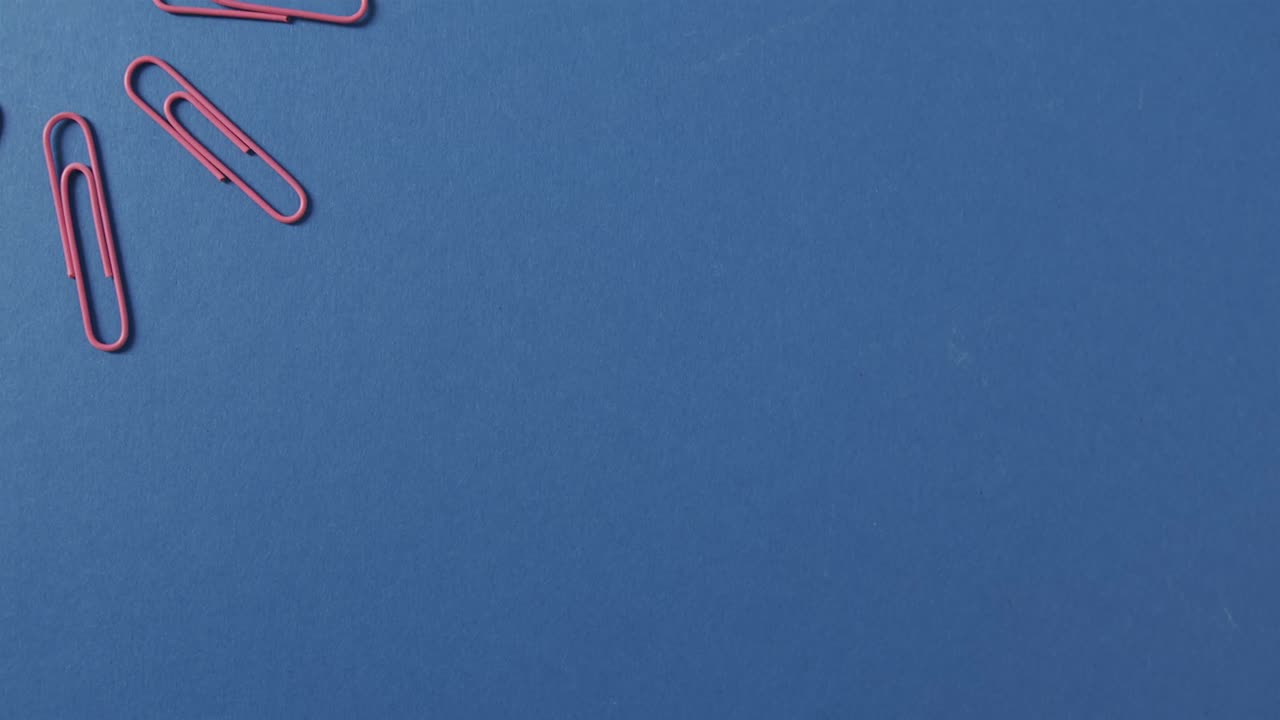 Vista dall'alto di graffette rosa disposte con spazio per il testo su sfondo blu, al rallentatore.
