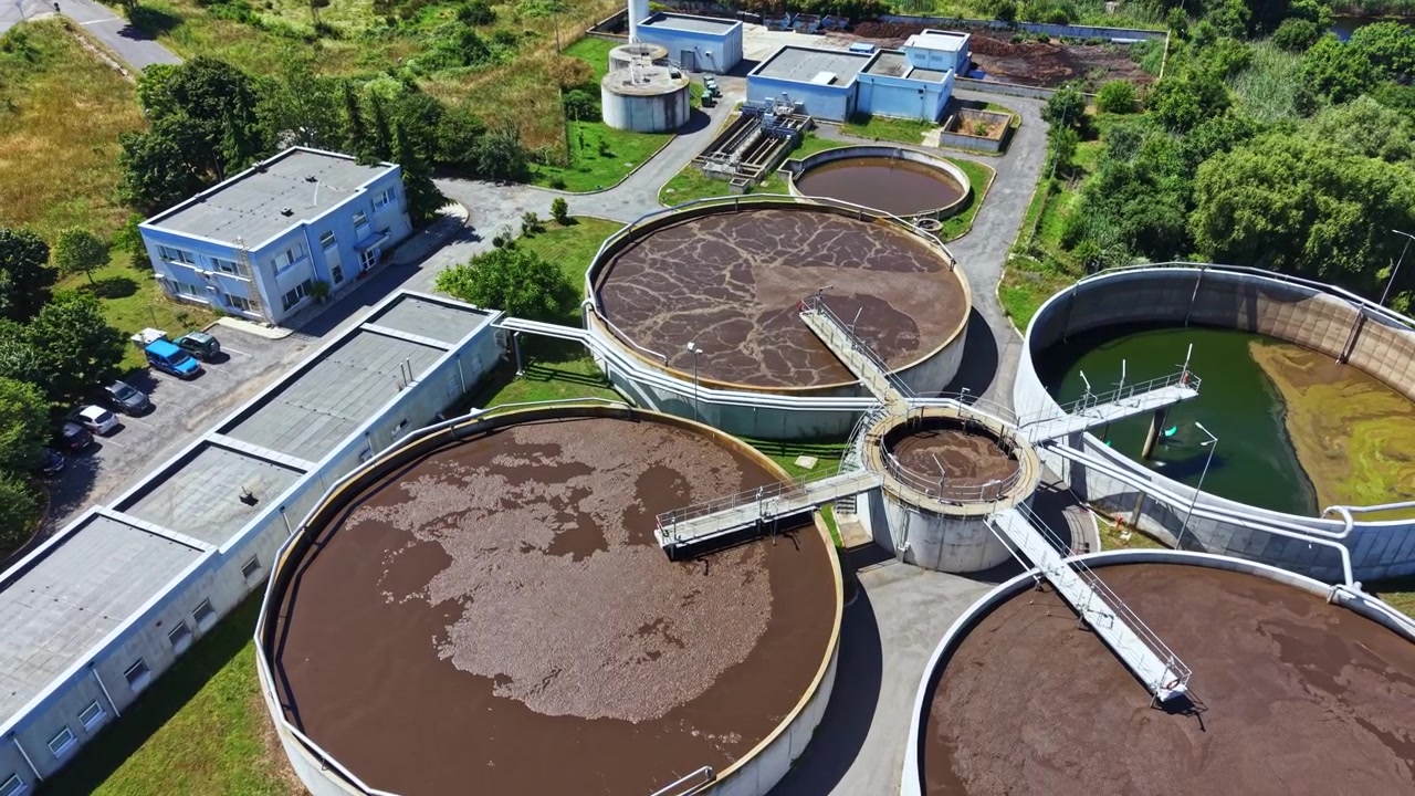 Planta de tratamiento de aguas residuales ubicada cerca de un río con múltiples tanques circulares y edificios que muestran actividad de procesamiento de aguas residuales durante el día.