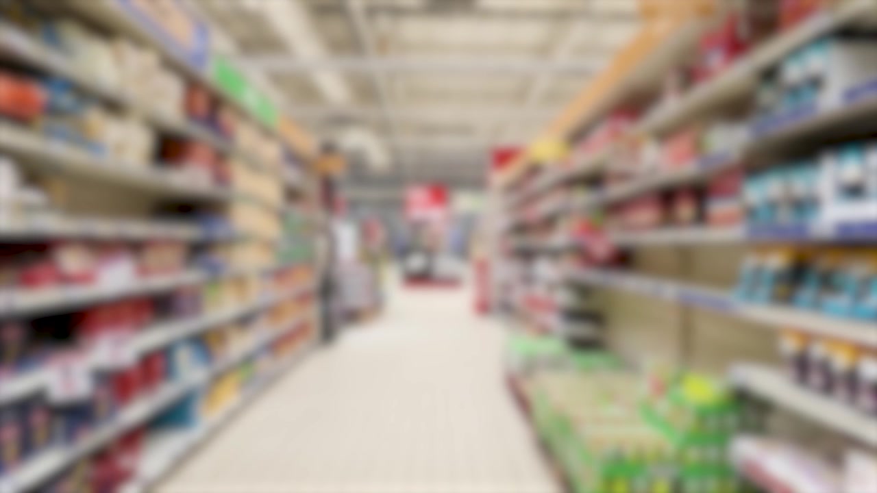 A blurred first-person perspective moves slowly down a bright supermarket aisle with stocked shelves