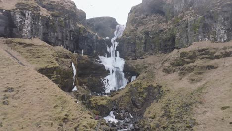 vista aérea de un hombre parado sobre una roca bajo una cascada helada, increíble paisaje de islandia, revelando un disparo de drone