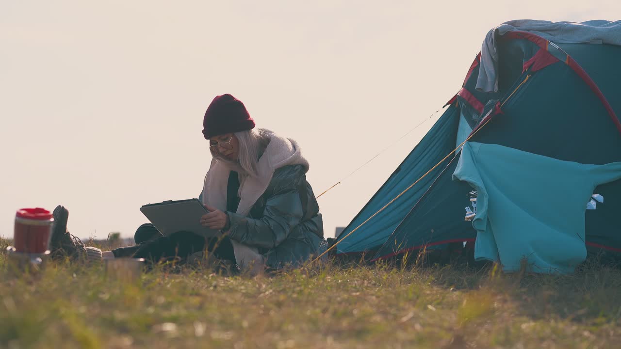 태블릿을 가진 여성 하이커가 가을의 초원에서 텐트에 앉아 있습니다.
