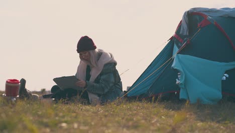 woman hiker with tablet sits at tent on autumn meadow