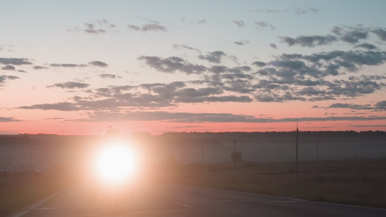 Motorcyclist wearing helmet riding countryside road at dusk with bright headlamp shining into lens, creating glowing flare effect against scenic sunset sky filled with drifting clouds