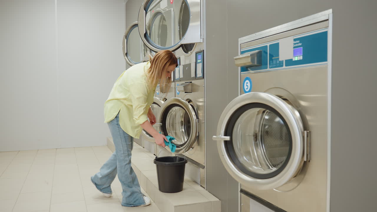 Industrial washing machines in row with open doors, stainless steel fronts, quiet laundromat scene as laundry worker walks toward units carrying cleaning bucket on tiled floor, maintenance concept