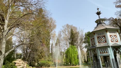 Wir-Sehen-Den-Chinescos-Teich-Im-Jardin-De-El-Principe,-Mit-Fokus-Auf-Das-Markante-Vogelhaus,-Den-Blauen-Himmel,-Die-Wasserstrahlen-Und-Alle-Baumkronen,-Aranjuez,-Spanien