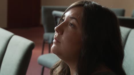 a serious close up of a pretty young ethnic female staring sadly at the altar inside a church sanctuary while sitting in a pew