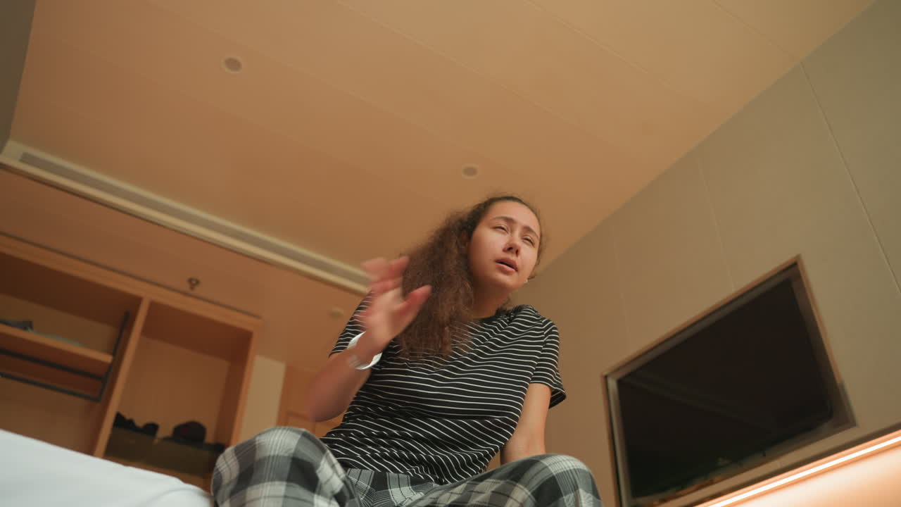Sitting on hotel bed, restless woman shifting to stand in plaid pajama pants, gestures toward closet and suitcase, interior shows television, open wardrobe and warm bedside lighting