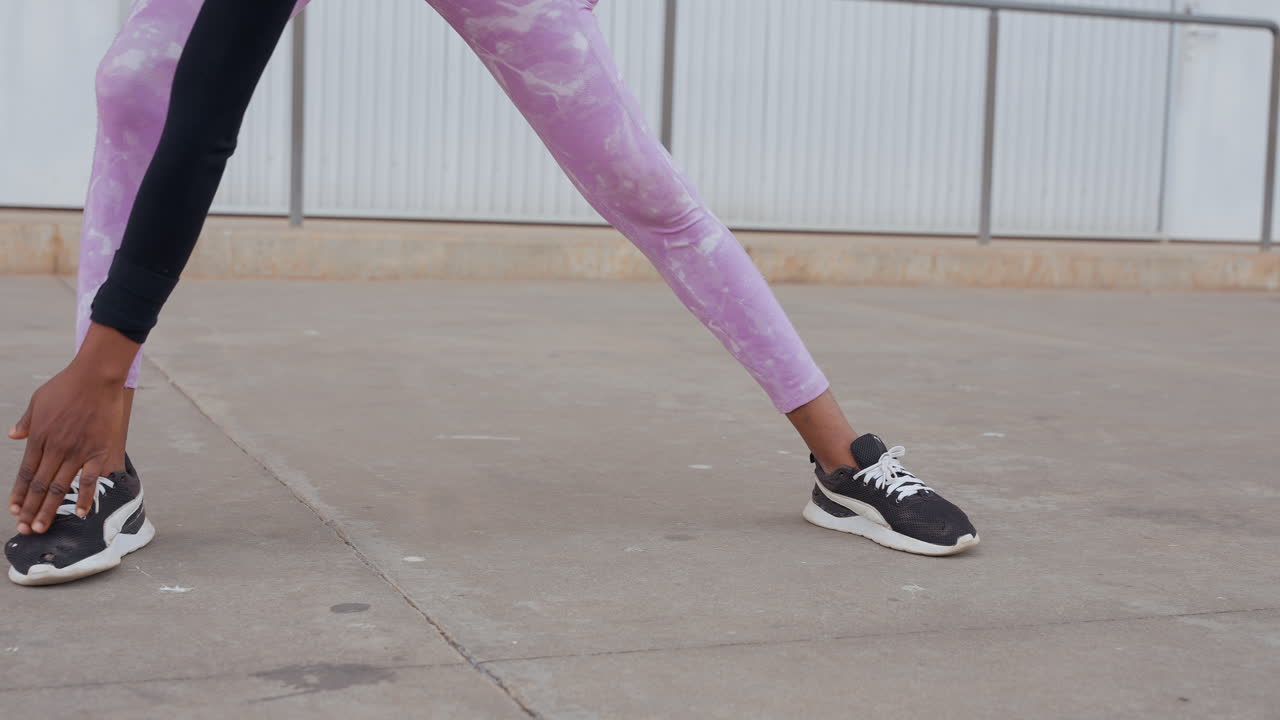 Donna di colore in allungamento a gambe larghe sul cemento, leggings viola e sneakers nere, ballerina urbana che si riscalda con un allungamento laterale e la mano che tocca il pavimento, sullo sfondo un lotto industriale vuoto, mobilità diurna.