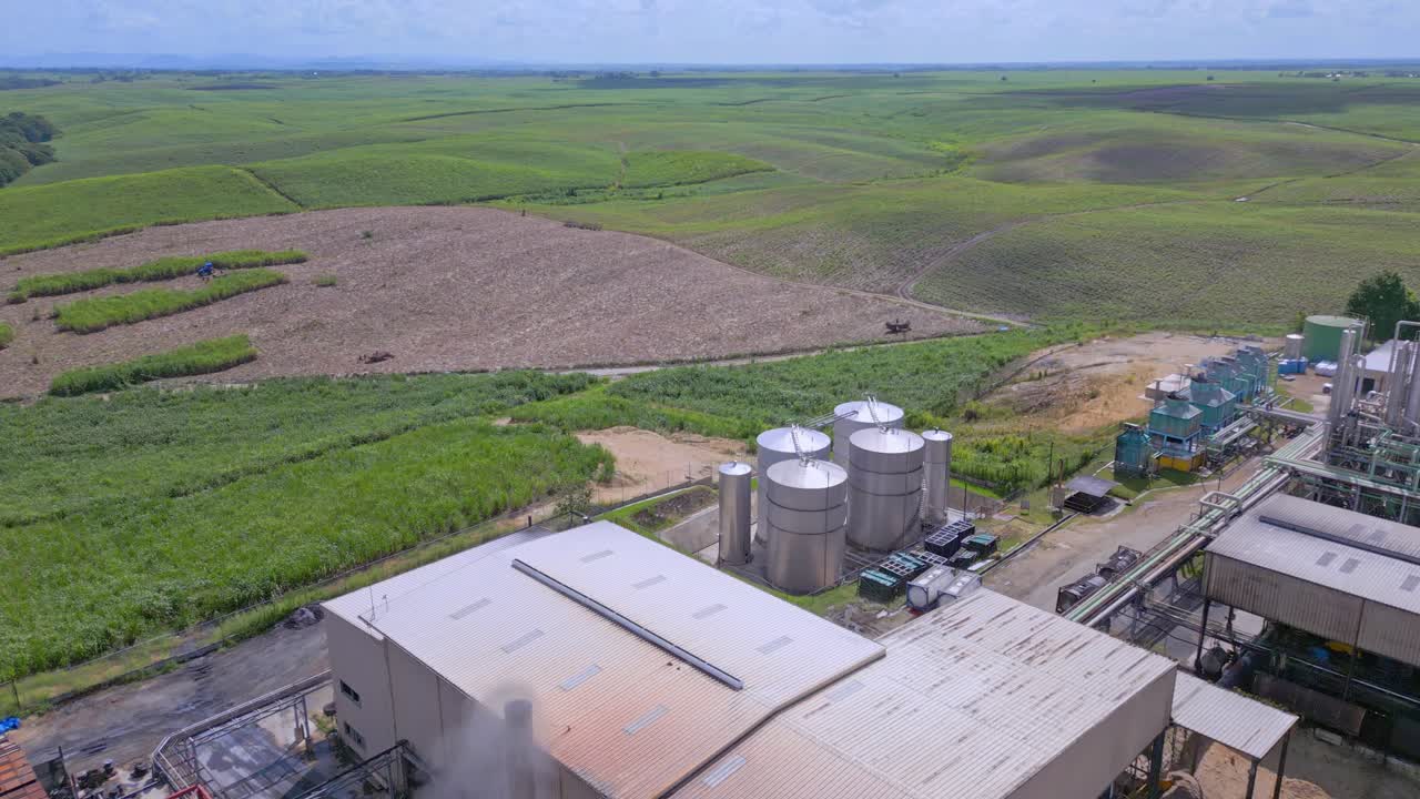 sobrevuelo aéreo antigua fábrica de ron con humos ascendentes en el cielo junto a campos de caña de azúcar en república dominicana