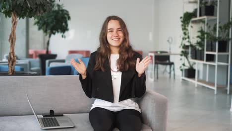 cheerful-girl-with-a-laptop-sits-on-the-couch,-emotionally-and-cheerfully-chatting-and-gesticulating-while-looking-into-the-camera-with-the-office-in-the-background