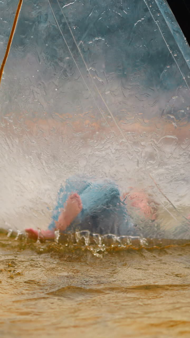 Little girl pushes legs lying on back inside large transparent water ball floating in small pond on gloomy day close view slow motion