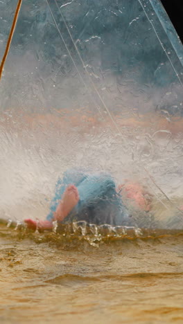 little girl pushes legs lying on back inside large transparent water ball floating in small pond on gloomy day close view slow motion