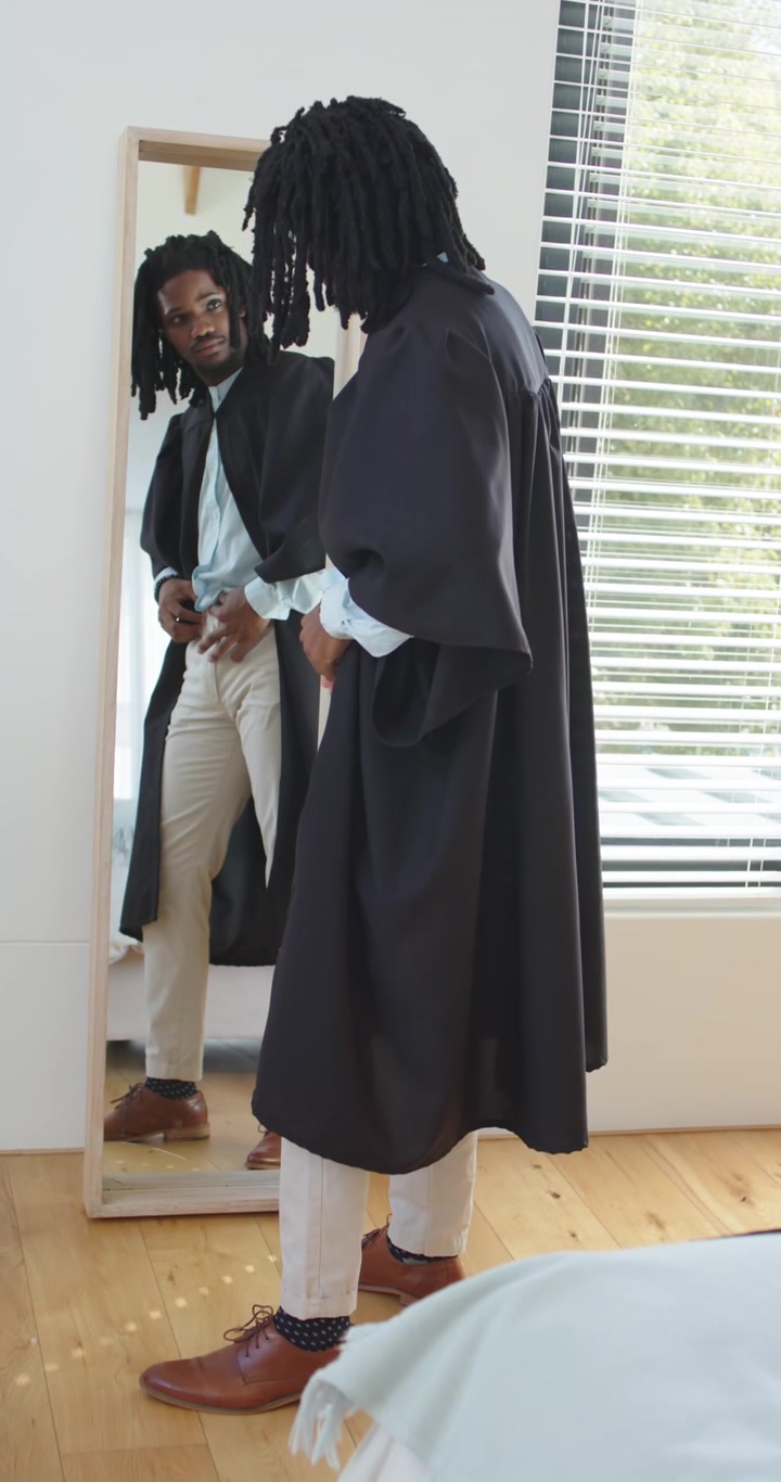 Vertical video: Adult African man adjusting robe tucking shirt, pants while checking fit in mirror