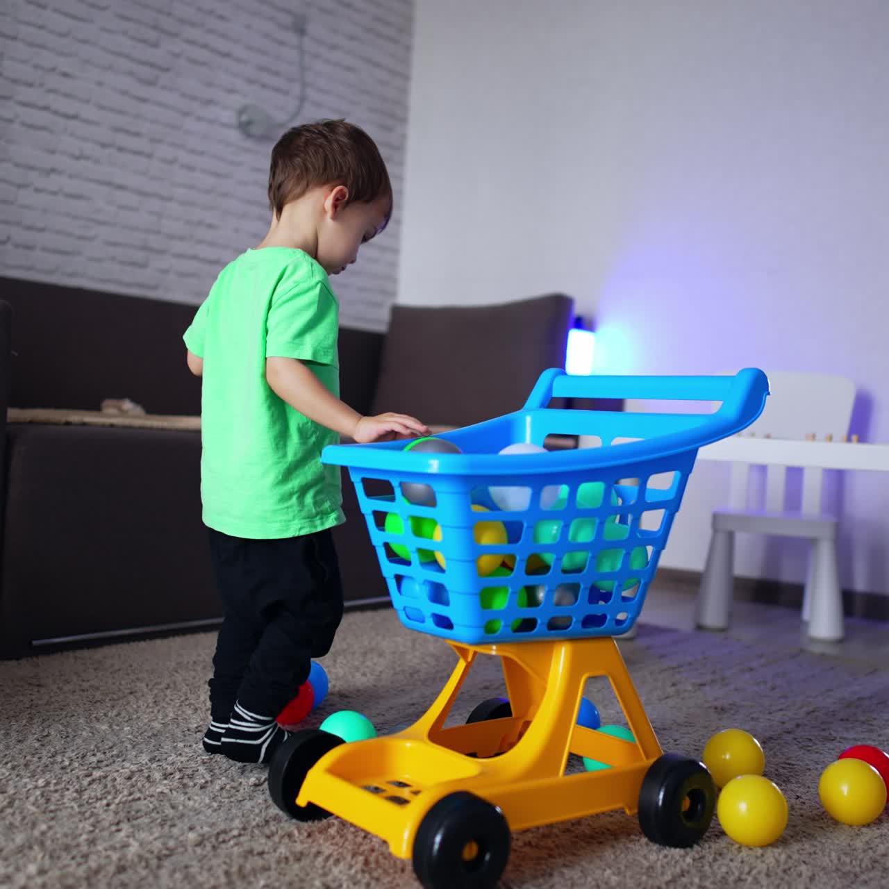 Um bebê caucasiano pega as bolas do chão e as coloca no carrinho. Um gato preto caminha ao redor da criança