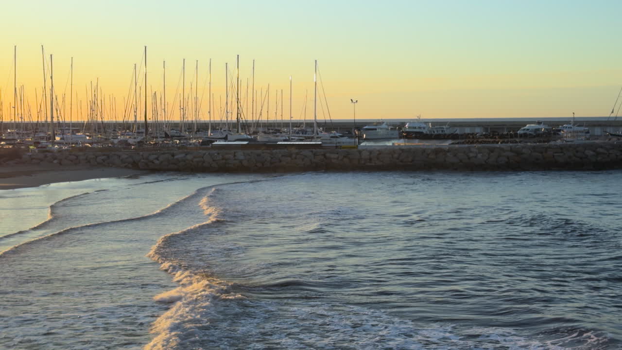 Tranquil sea waves roll toward the shore at sunset with a scenic marina in the background. Dozens of moored sailboats, golden reflections, and a peaceful coastal mood