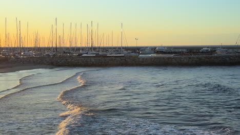 tranquil sea waves roll toward the shore at sunset with a scenic marina in the background. dozens of moored sailboats, golden reflections, and a peaceful coastal mood