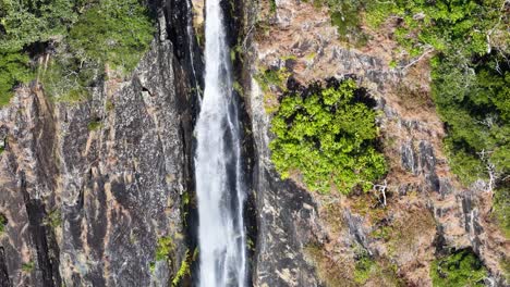 Drone captures a stunning vertical waterfall cascading through dense greenery and rocky cliffs in a vibrant rainforest setting