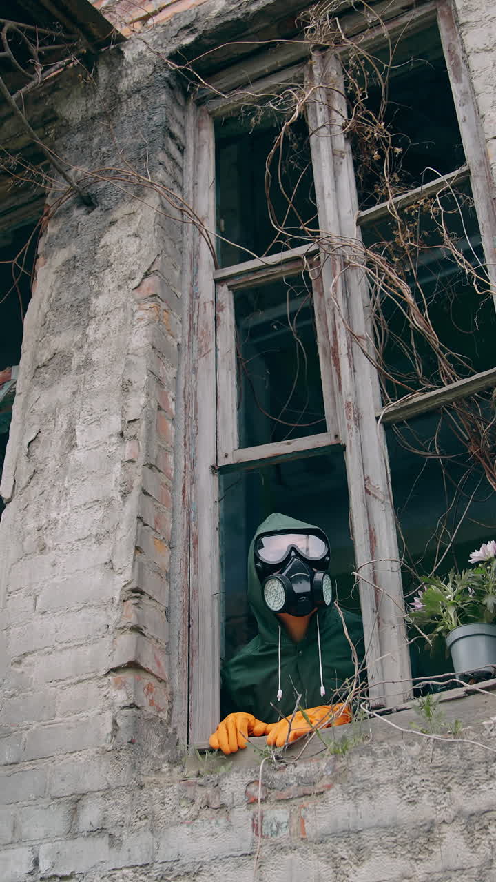 Ecologist inside the ruined building. Scientist in safety gas mask looking out of the window from abandoned building. Flowers in flower pot in window sill. Vertical video