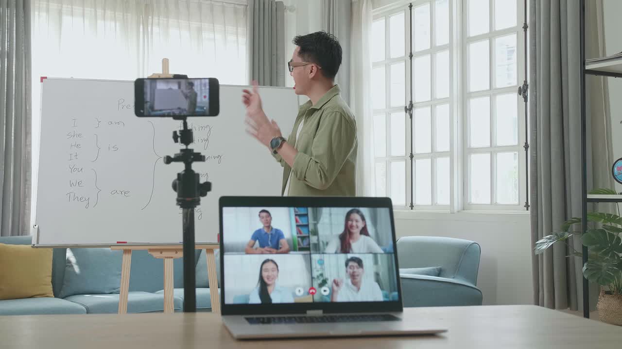 Asian Male Teacher With Glasses Shoots Video By Smartphone Camera And Having Video Call On Laptop While Teaching English At Home