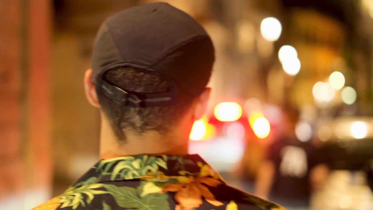 Slow Motion Close Crop Gimbal Rear Following View of Young Tan Man with Black Hat and Hawaiin Shirt Walking Through Crowded Street at Night