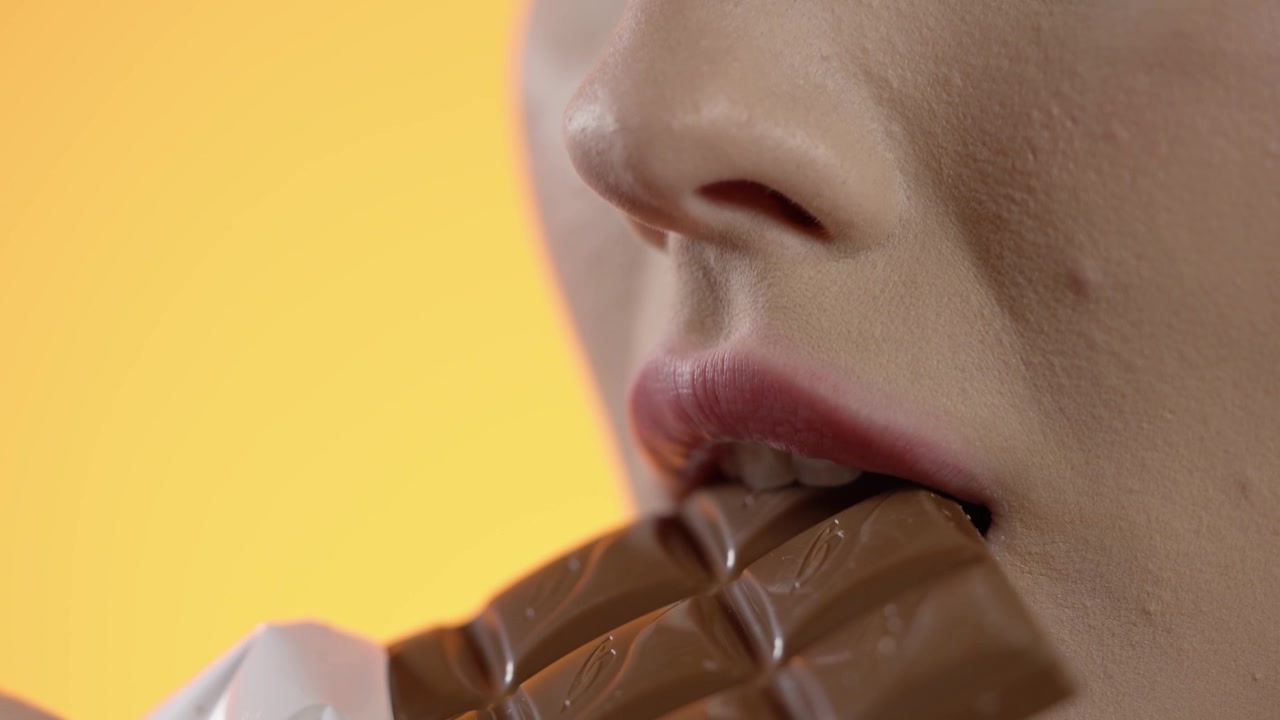 woman taking a bite of a chocolate bar, close-up biting of snack on pink background