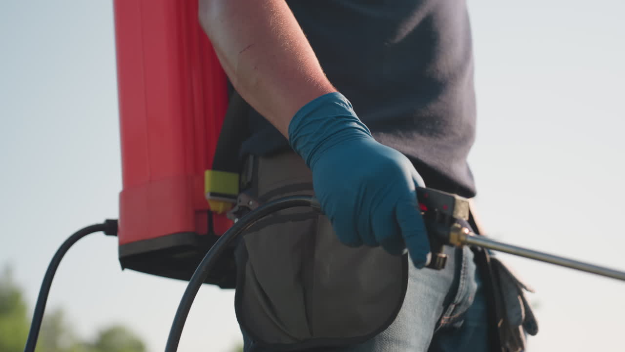close up of man fumigating field with backpack sprayer wearing glove and protective gear, sunlight highlighting arm and hose during outdoor agriculture pest control