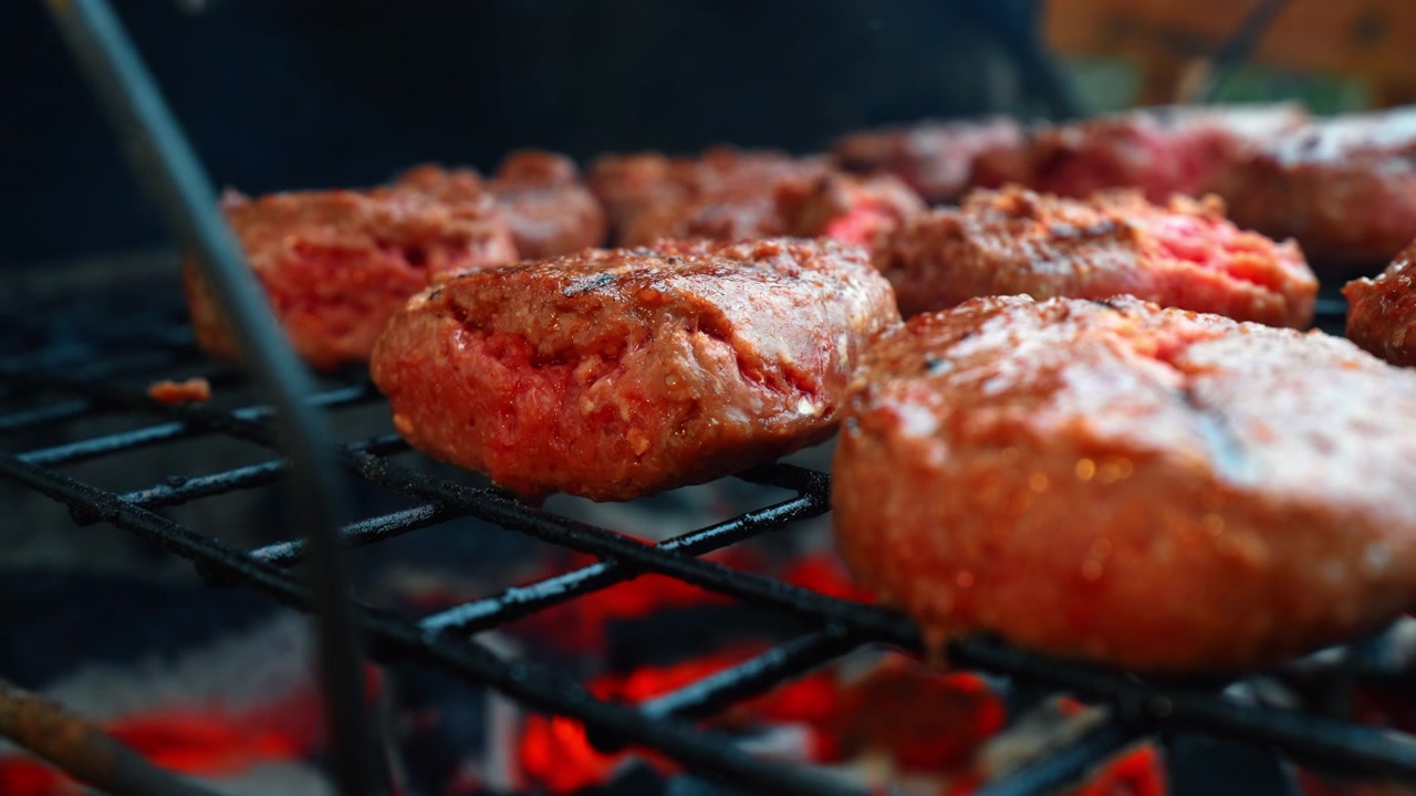 Hamburger di manzo crudi che cuociono su una griglia a carbone. Primo piano di polpette di carne rossa su una griglia per barbecue con braci ardenti sotto.