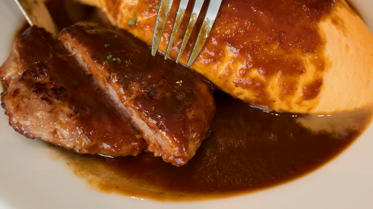 Close-up of fork slicing juicy hamburg steak beside omelette rice, rich brown sauce, warm lighting
