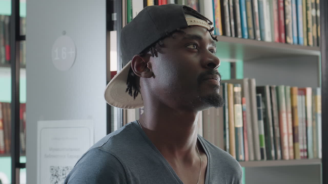 Close up of young man in casual cap turning head with elegant smile in bright library, standing near shelf filled with books, exuding confident relaxed mood with subtle expression