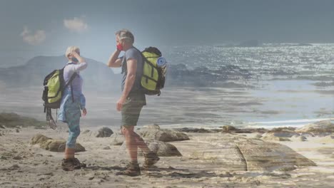 happy caucasian senior couple wearing facemasks hiking by the coast over fast moving clouds