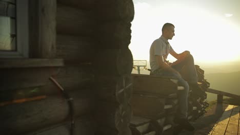 young man sit near old wooden house. sun beams. slow motion