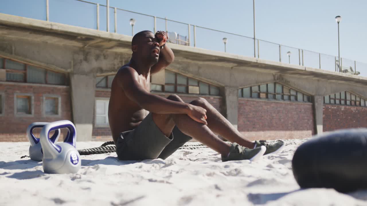 Træt afroamerikansk mand sidder, tørrer sin pande og holder pause under udendørs træning på stranden.