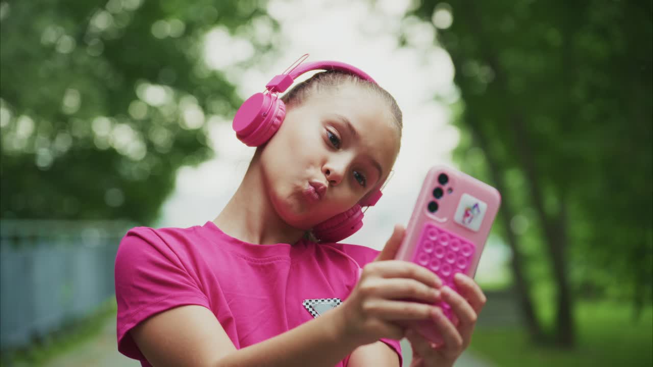 A Young Girl in Pink Headphones Captures Joyful Moments with Her Phone While Enjoying a Lush Greenery Background, Exuding Confidence and Playfulness