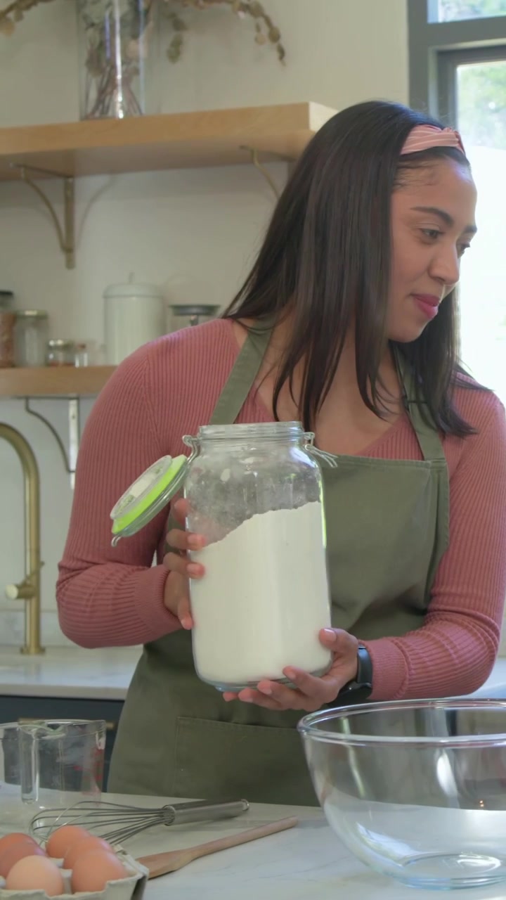 Vídeo vertical: Mulher de avental verde, inclinada, adicionando farinha de um pote para assar no balcão com uma criança.